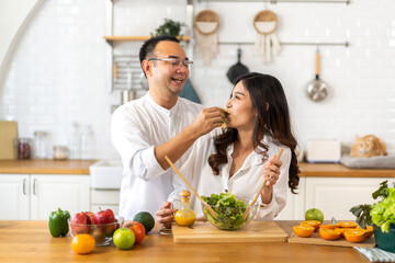Young asian family couple having fun cooking and preparing cook vegan food healthy eat with fresh vegetable salad on counter in kitchen at home.Happy couple looking to preparing food