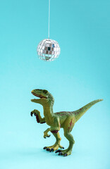 Fototapeta premium Happy cute green toy dinosaur dancing under disco ball on blue background. Minimal creative art poster.