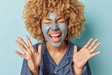 Emotional curly haired woman applies facial mud clay mask keeps palms raised up exclaims loudly dressed in pajama undegoe beauty procedures at home after awakening isolated over blue background