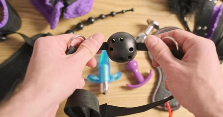 Male hands inspect a bdsm ball gag for erotic games. POV view, a bunch of toys from the sex shop are on the table.