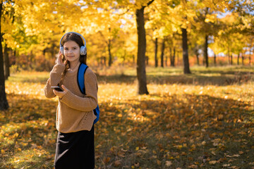 Caucasian young girl teenager schoolgirl pupil using smartphone while listening to the music in headphones going back to school in autumn park.