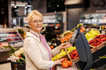 A cheerful old woman is measuring tomatoes on scales at the supermarket and smiling at the camera.