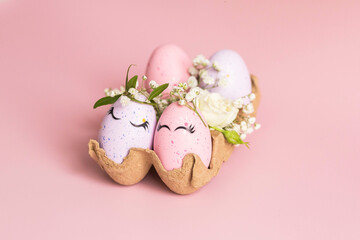 Stylish Easter pink and liliac eggs with cute faces in floral wreath crowns in carton tray on pink background. Easter decorations and gift. Happy Easter concept. Copy space. Isolated.