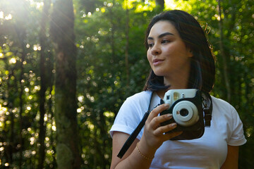 Mulher jovem com cabelo escuro segurando uma câmera de foto instantanea no meio da mata em um dia...
