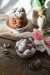 Bowl with white, pink and foiled chocolate eggs, homemade Easter sweet bread, pink tulips and feathers on rustic wooden table.