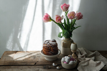 Homemade Easter bread (cruffin), chocolate eggs, wax egg-shaped candle and pink tulips on old wooden table in the evening sunlight.