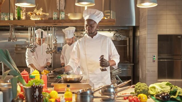 Portrait of African American professional skillful chef frying vegetables on pan and mixing. Experienced successful male cook cooking at modern kitchen and wearing white uniform. Flames