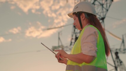 Young woman engineer, power engineer in helmet checks power line using computer tablet online. Electrician in outdoors. Electric lines of high voltage at sunset. Distribution and supply of electricity