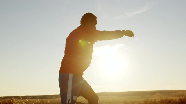 African-american man in boxing gloves practicing boxing against background sky outdoors, side view. Black man trains martial arts against background of sun. Martial art concept