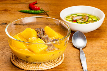 Traditional delicious homemade potato, chilli, beef onion and coconut milk curry in transparent glass bowl on bamboo mat with slice cucumber, onion, vinegar and chilli sauce in white ceramic cup on wo