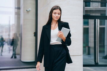 Pensive Asian brunette young businesswoman in black suit against building holds laptop looks at...