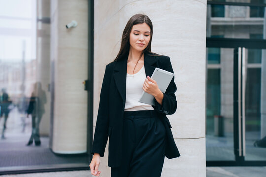 Pensive Asian Brunette Young Businesswoman In Black Suit Against Building Holds Laptop Looks At Camera. Attractive Female Entrepreneur Planning Day. Purposeful Student Girl Preparing For Exam.
