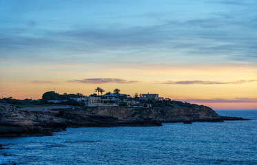 A stunning mansion perched on a mountain at the shore, surrounded by the sea. Palm trees, rocks, and the beautiful colors of the sunset dominate the landscape