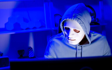 Male hacker wearing mask and hoodie, sitting in dark room, looking at camera, making cyber attack criminal to privacy password, hacking money, currency system in internet networking.