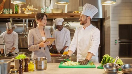 Portrait of attractive professional caucasian chef chopping vegetables and talking with beautiful cute female manager with tablet. Two colleagues discussing work at kitchen of restaurant.