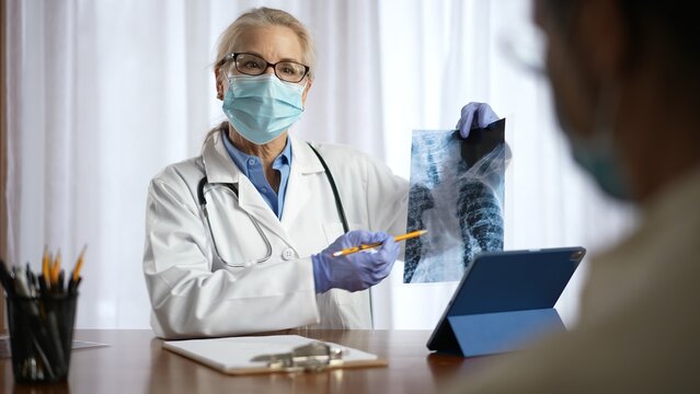 Woman Doctor Wearing Gloves And Face Mask Shows X-ray To Patient. Concept Of Medical Visit During Pandemic.