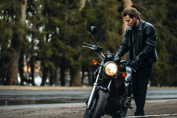 male motorcyclist in a leather motorcycle jacket stands by a motorcycle on a forest road....