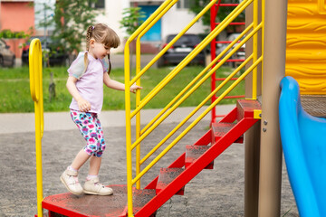 Cute little girl climbs the steps of the slide on the playground, side view. A happy pretty little girl with long pigtails is playing on an outdoor playground. Active children's outdoor games