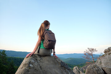 Woman hiker seated alone on rocky mountain cliff enjoying view of evening nature on wilderness trail. Active way of life concept