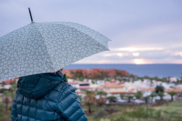Back view of senior woman in a rainy evening holding an umbrella while strolling outside. Horizon over water. Copy space