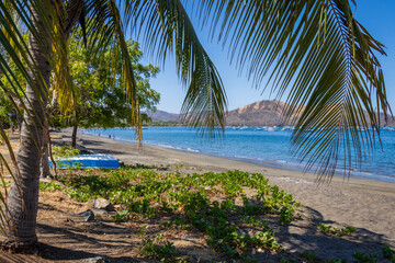 An image of a tropical beach with gentle waves rolling into a sand beach. Off in the distance are hills and anchored boats. Framing the top of the image are palm tree branches. 