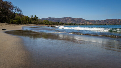 A peaceful tropical image of waves crashing onto a smooth sand beach in Costa Rica. In the distance are hills and palm trees. The beach is mainly deserted of people. 
