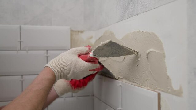 The master glues white tiles. A tile laying master takes a mixture for gluing finishing materials on a wall, close-up. Stylish trendy white chamfered ceramic tile on the kitchen wall.