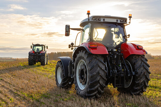Fototapeta Back view of tractors driving on field at sunset. Tractors riding to gather crop of wheat. Bright light of sunset illuminates rural environment. Scenic view of countryside at background.
