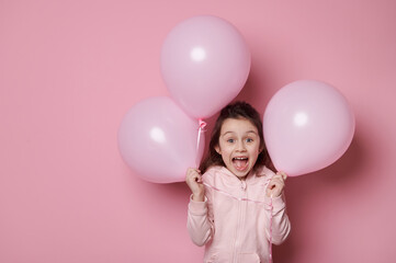 Fototapeta na wymiar Amazed little child girl in pink hoodie, holding pink pastel balloons, expressing surprise and amazement, posing with open mouth on isolated pink color background with copy advertising space for text