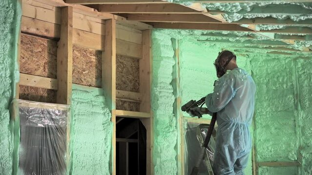 Male builder insulating wooden frame house. Man worker spraying polyurethane foam inside of future cottage, using plural component gun. Construction and insulation concept.
