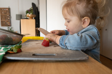 Blonde toddler learning to cut soft fruits with a safety knife; working to combat picky eating by getting child involved in preparing food