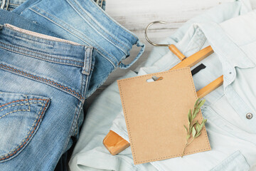 Sustainable still life with Stack of blue jeans and denim shirt on hanger and empty craft card. Second hand apparel idea. Circular fashion, donation, charity concept - obrazy, fototapety, plakaty
