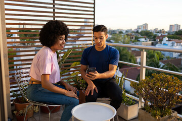 Two Latin friends sitting on the balcony looking at the mobile phone with a view of the city in the background