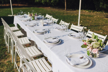 Fototapeta na wymiar A large white long table with chairs, decorated with fresh flowers, with plates, glasses and forks, stands in a park with grass. Wedding decorations and details. Preparing for a wedding party.