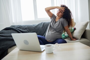 Obraz premium Pregnant woman with headache and fatigue sits at home on sofa with a laptop and watches tv shows movies and series. Lifestyle of a pregnant woman, preparation for childbirth, last month of pregnancy
