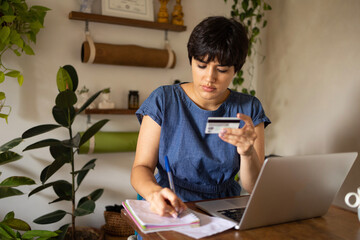 Young latin girl writing while holding credit card. She is sitting with a laptop in apartment full of plants