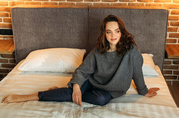An attractive woman is sitting on a bed in a bedroom wearing blue trousers and a gray jumper.