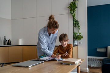 Young Caucasian mother helping child son with homework at home, sitting together at kitchen table....
