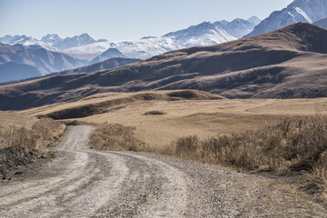 Road in the autumn mountains against the backdrop of a rocky ridge of mountain peaks with the first snow, sunny autumn day in the mountains landscape