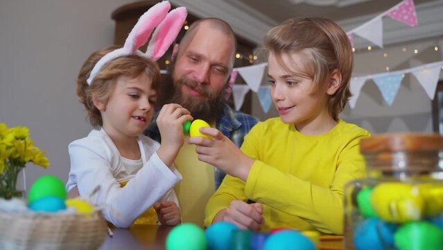 Easter Family traditions. Father and two caucasian happy children with bunny ears playing with Easter eggs with paints for holidays while sitting together at home table. Kids embrace and smile in cozy