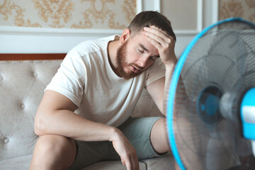 Young bearded man using electric fan at home, sitting on couch cooling off during hot weather,...