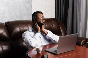 African man feels neck pain after working for long hours with laptop online in office. Cervical spine osteochondrosis is radicular syndromes of african american man.