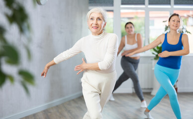Cheerful sporty senior woman training movements of rhythmic latin zumba with group in fitness...