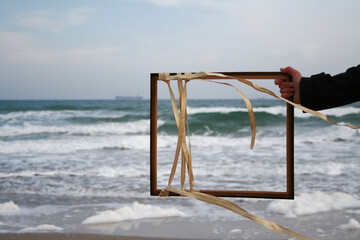 A girl against the sea holds a frame with ribbons