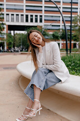 Fototapeta premium Full-length photo of beautiful relaxed girl wearing white jacket and jeans and heels is sitting outdoor on background of business district and resting with lovely smile and closed eyes. 