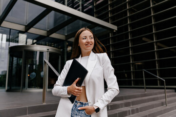 Fototapeta premium Close up outdoor photo of smiling adorable woman is wearing white jacket and top is looking aside, holding laptop and posing over business district in city