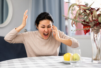 Fototapeta na wymiar Emotional Asian woman sitting at a table in an apartment swears at someone while talking on a mobile phone
