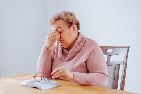 Depressed Senior Woman at Home Struggling with Eye Pain While Reading. A senior citizen sits at home with a book in her hand, her glasses perched on her nose as she struggles to read.