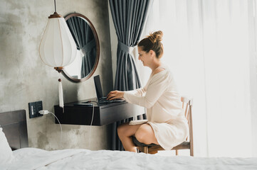 Side view full body of young woman in white robe sitting on chair at wall mounted table and typing text on netbook while surfing internet during weekend in bedroom