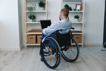 A man in a wheelchair freelancer works at a laptop at home, restriction of movement, view from the back, copy space, integration into society, health concept a person with disabilities, a real person